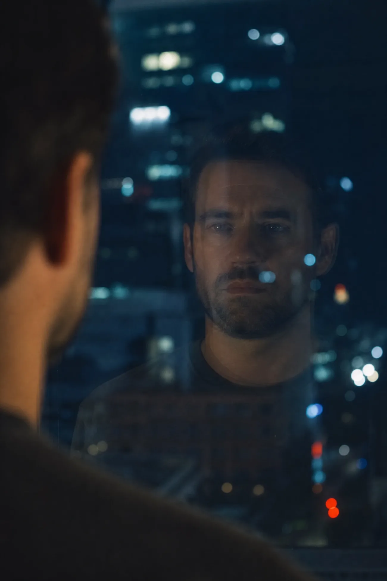 Un visage et son reflet légèrement décalé dans une vitre de bureau, la nuit.